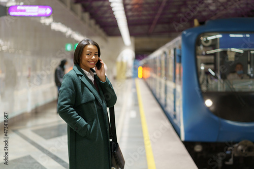 Happy successful businesswoman returning home from office talk on smartphone wait for train at subway station. Smiling african woman call to family or husband from underground after long working day