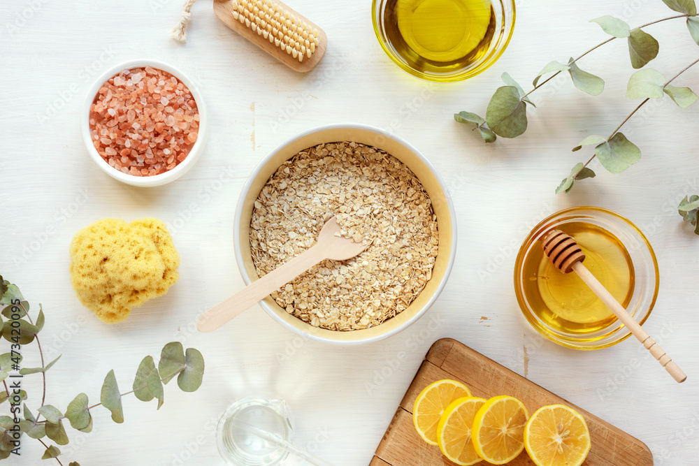 Natural ingredients for homemade scrub. Oat flakes, oil, honey, pink himalayan salt, lemon, face or body brush, sponge and eucalyptus branches on white wooden table. Top view, flat lay Natural ingredients for homemade scrub. Oat flakes, oil, honey, pink himalayan salt, lemon, face or body brush, sponge and eucalyptus branches on white wooden table. Top view, flat lay