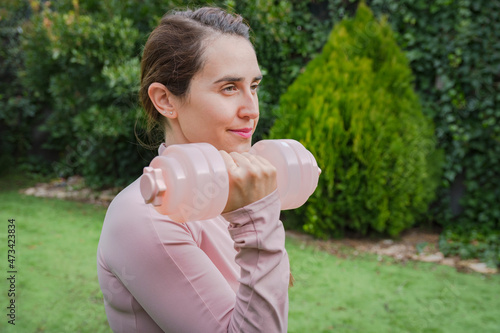 Woman dressed in pink lifting dumbbell in yard, home fitness photo