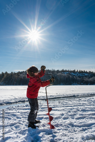 Young boy using auger to make a hole in the ice on a sunny winter day.