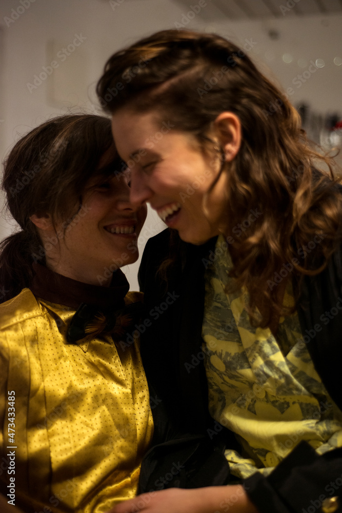 queer lesbian couple laugh at home in kitchen armchair at dinner party queer lesbian couple laugh at home in kitchen armchair at dinner party