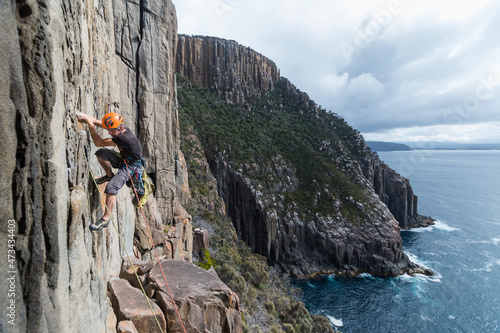 Male rockclimber climbs the exposed edge of a dolerite sea cliff using two ropes as protection in a cloudy day, with bushy cliffs and the ocean in the background in Cape Raoul, Tasmania photo