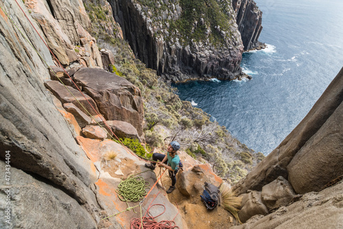 Woman belaying on the exposed edges of a dolerite sea cliff using two ropes as protection in a cloudy day, with bushy cliffs and the ocean in the background in Cape Raoul, Tasmania, Australia photo