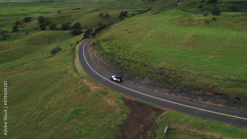 Aerial tracking shot of Jeep driving on curvy road in Hawaii with lush green landscape.