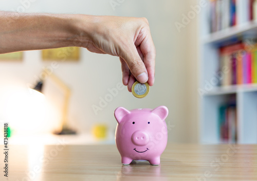 Hand putting 1 Real (Brazilian currency) coin into a piggy bank, with a house interior in the background of the image.