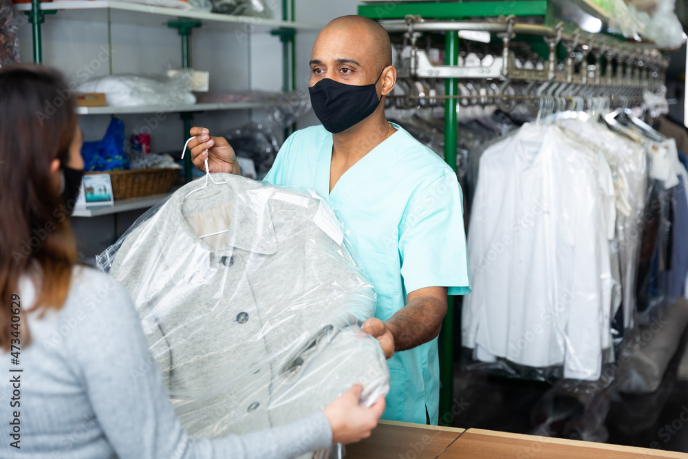 Positive man laundry worker in protective mask for disease prevention returning clean clothes to customer at dry-cleaning facility