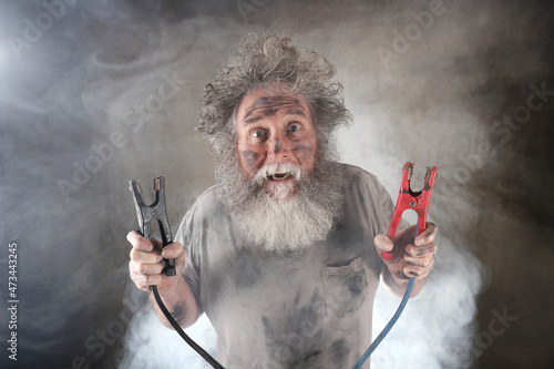 Crazy old man with beard holding jumper cables surrounded by smoke receiving a shock photo