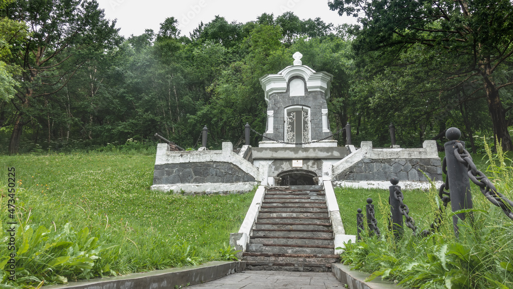 The old gray-and-white chapel stands in a park on a hill. The latticed doors are open. A stone staircase leads to the entrance. There is a green lawn around, lush vegetation. Petropavlovsk-Kamchatsky.