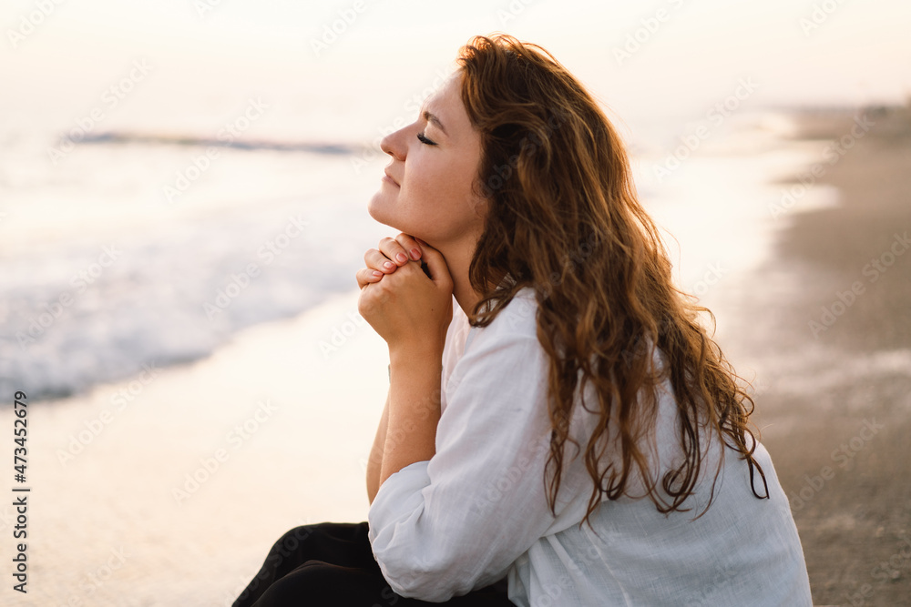 Woman closed her eyes, praying on a sea during beautiful sunset. Hands folded in prayer concept for faith, spirituality and religion. Peace, hope, dreams concept - obrazy, fototapety, plakaty 
