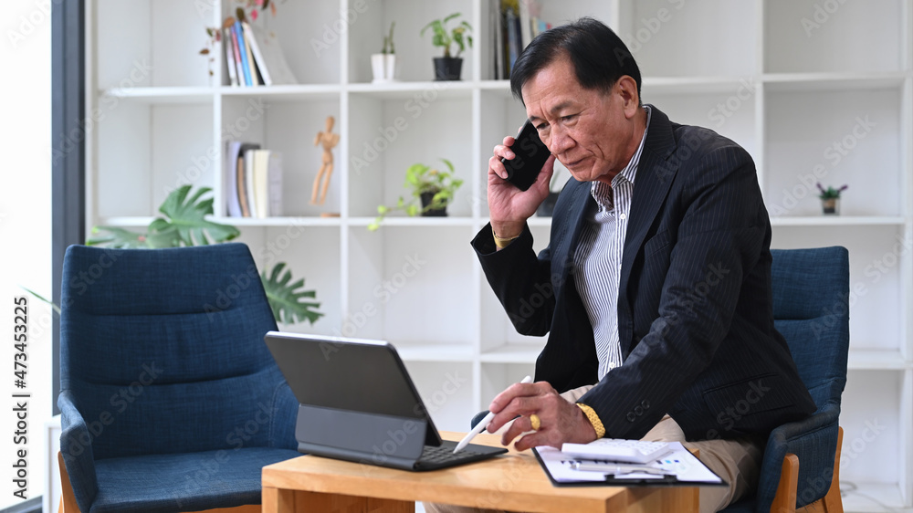 Mature businessman talking on mobile phone  and working with computer tablet in his office room.