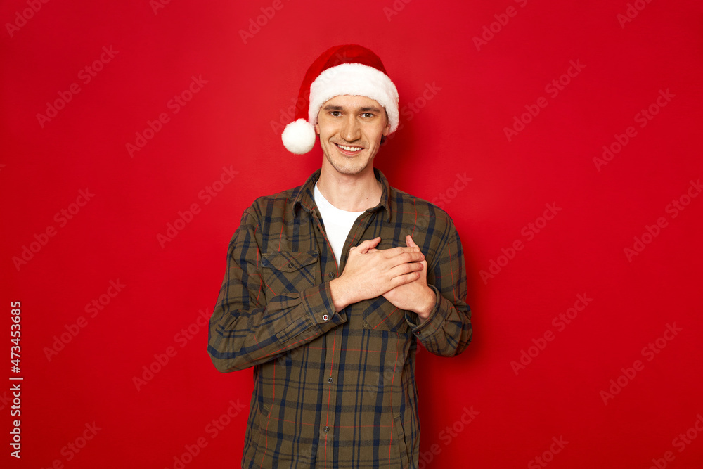 Indoor shot of handsome man keeps hands on chest, being cordial and friendly, expresses gratitude and thankfulness, has pleasant smile, dressed in casual outfit xmas hat, isolated on red studio wall Indoor shot of handsome man keeps hands on chest, being cordial and friendly, expresses gratitude and thankfulness, has pleasant smile, dressed in casual outfit xmas hat, isolated on red studio wall