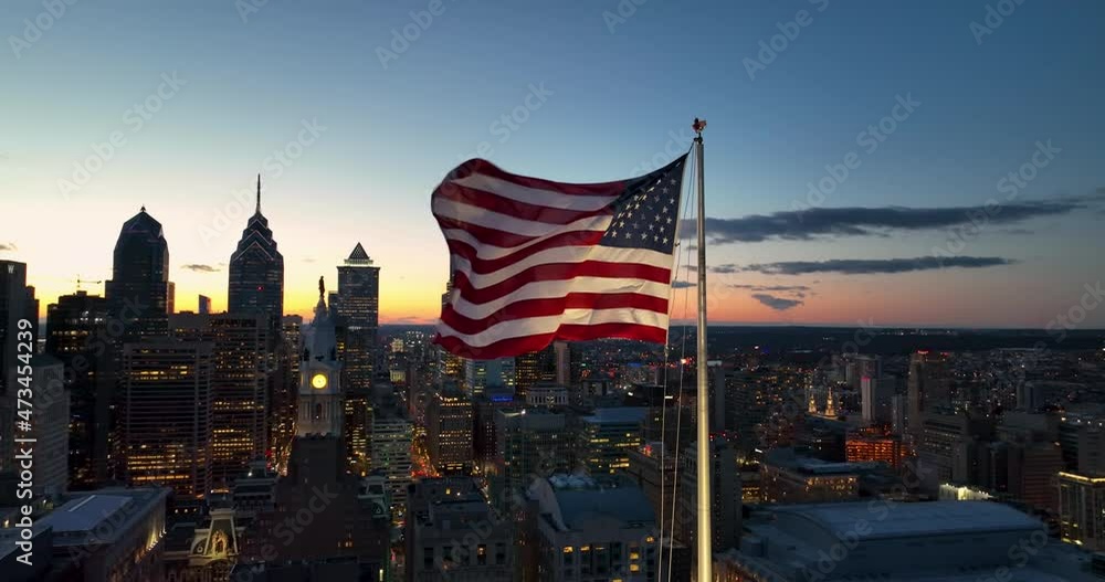 American flag waves in urban city USA. Skyscraper silhouette at sunset. Cinematic powerful shot.