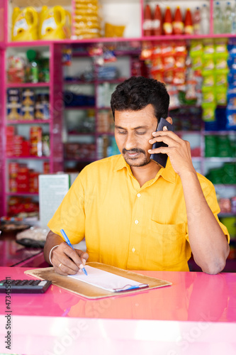 Kirana marchant or owner taking orders by noting from customer over Mobile phone call at groceries shop - concept of small business and Home delivery services photo