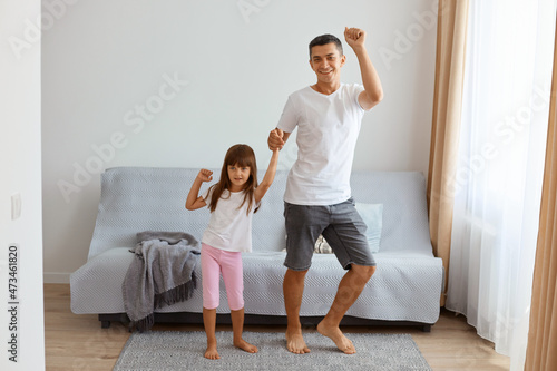 Full length portrait of attractive male wearing white t shirt and jeans short dancing with his daughter indoor near sofa in living room, expressing positive emotions. photo