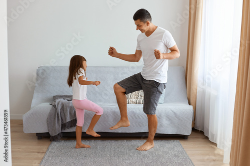 Excited extremely happy father wearing jeans short and white t shirt dancing with his daughter against sofa in living room, raising their legs, having great time together. photo