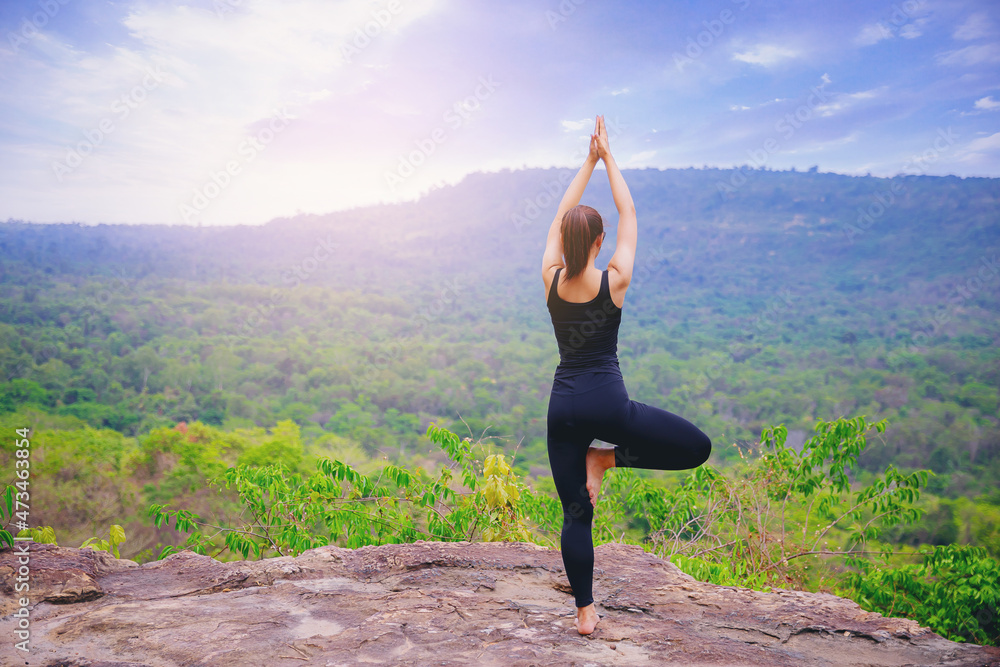 Young woman standing and doing yoga pose at the top of mountain in the morning