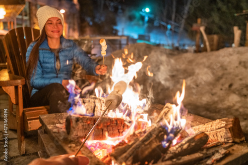 Winter campfire couple roasting marshmallows in firepit for smores at ski holiday resort. Asian happy woman relaxing after ski.