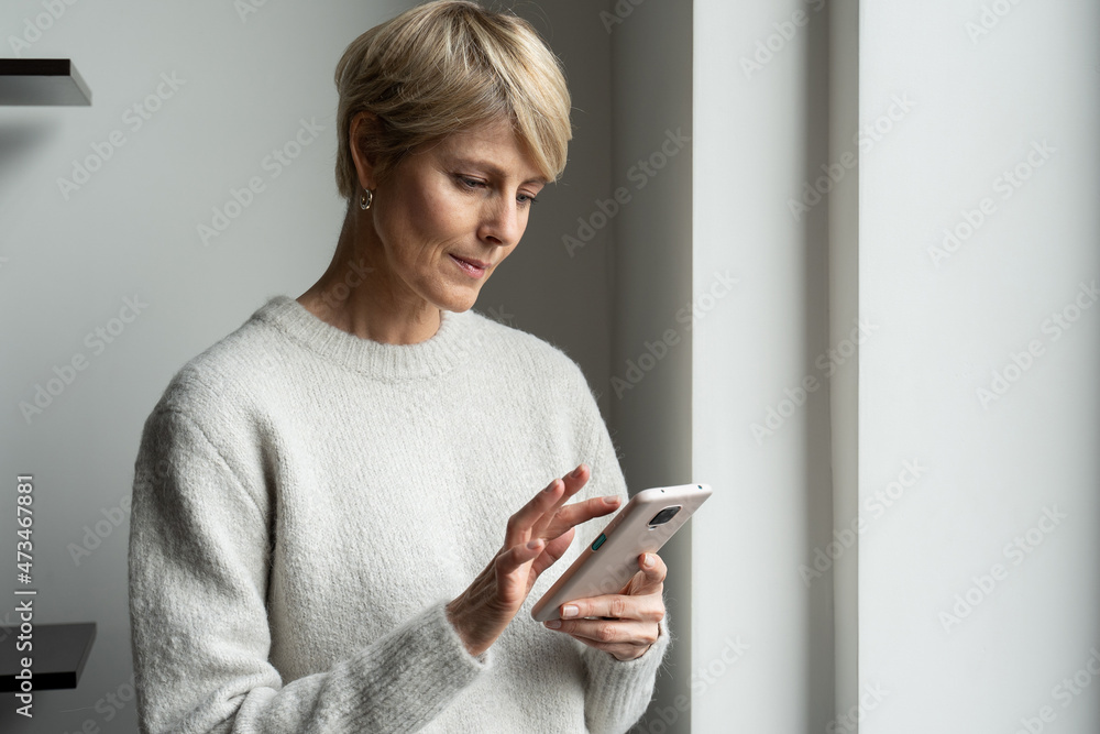 Charming middle-aged woman standing at the window and typing text messages on her phone, using a modern smartphone on the Internet Charming middle-aged woman standing at the window and typing text messages on her phone, using a modern smartphone on the Internet