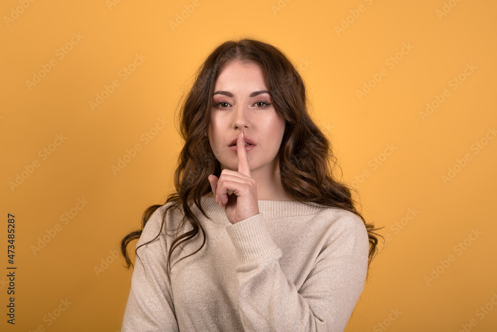 A beautiful and attractive Caucasian brunette girl with wavy hair in a casual jumper showing a sign of silence, a serious expression. Girl isolated on orange studio background. A beautiful and attractive Caucasian brunette girl with wavy hair in a casual jumper showing a sign of silence, a serious expression. Girl isolated on orange studio background.