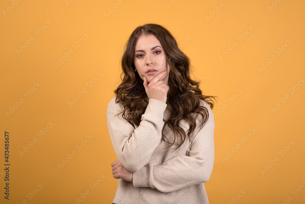 A young and attractive Caucasian brunette girl with wavy hair in a casual jumper holding her hand on her chin and thinking on an orange studio background. A young and attractive Caucasian brunette girl with wavy hair in a casual jumper holding her hand on her chin and thinking on an orange studio background.