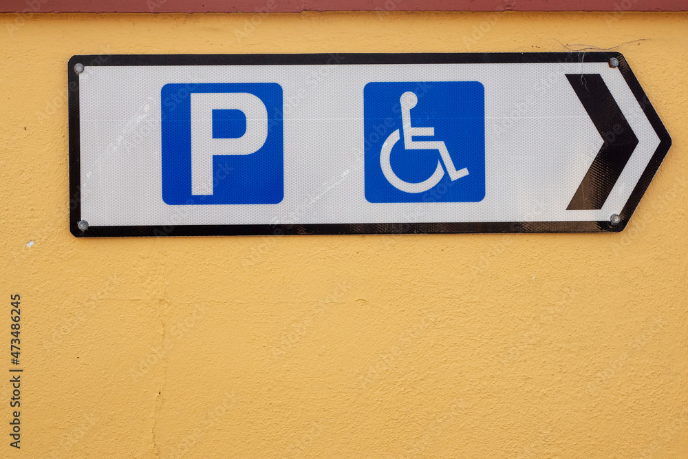 White sign on a yellow textured wall for parking with wheelchair access symbols. Copy space. Care for disabled people and members of their family. Blue and yellow complimentary color scheme. White sign on a yellow textured wall for parking with wheelchair access symbols. Copy space. Care for disabled people and members of their family. Blue and yellow complimentary color scheme.