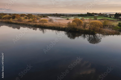 Nature scene. Calm water and trees reflection. Fog over meadow in the background. Calm and relaxing atmosphere. Soft pastel cloudy sky. View on a River Corrib, Galway city, Ireland.