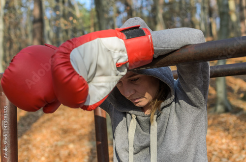Young charming woman, in a tracksuit with a hood on her head, in red boxing gloves, is resting after training in women's boxing in the autumn forest in nature