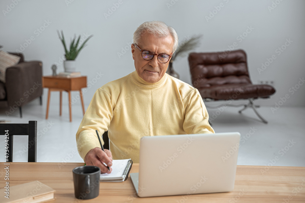 Senior man wearing glasses sitting in front of the laptop and writing notes Senior man wearing glasses sitting in front of the laptop and writing notes