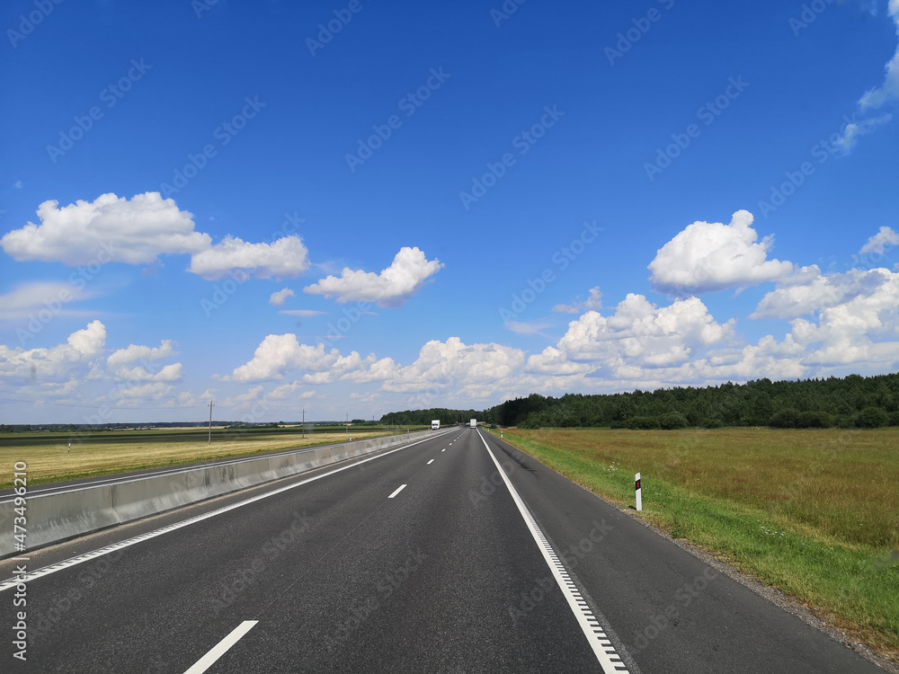The road is covered with asphalt under a blue sky with clouds, a white truck is coming towards us