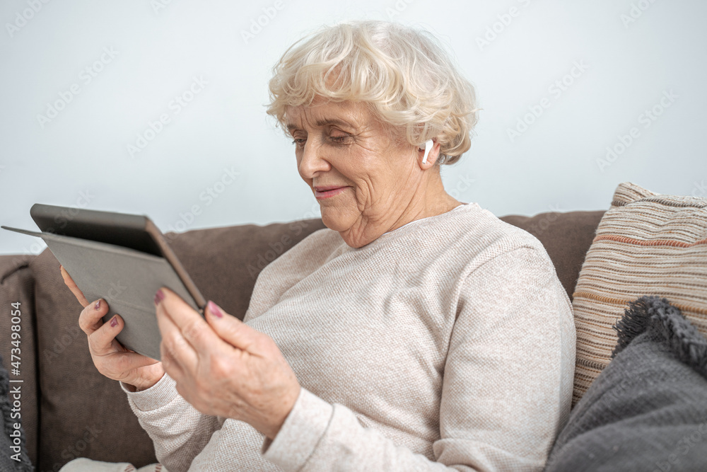 Grandmother wearing wireless earphones sitting on couch in living room and using tablet Grandmother wearing wireless earphones sitting on couch in living room and using tablet