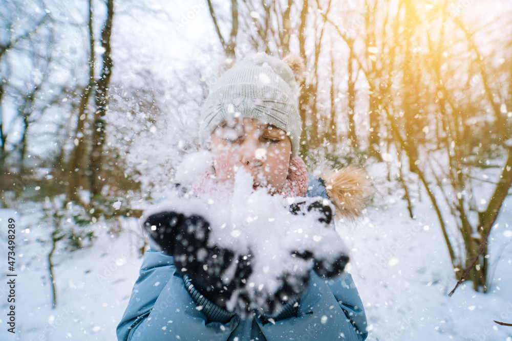 Fototapeta premium Girl playing on snow in winter time. Happy children in beautiful snowy winter forest on Christmas day. girl blows snow on snowflakes bokeh background. Happy childhood, active winter holidays concept 
