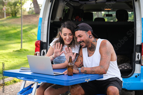 Young tattooed couple greet and talk in a laptop video call during a van trip.