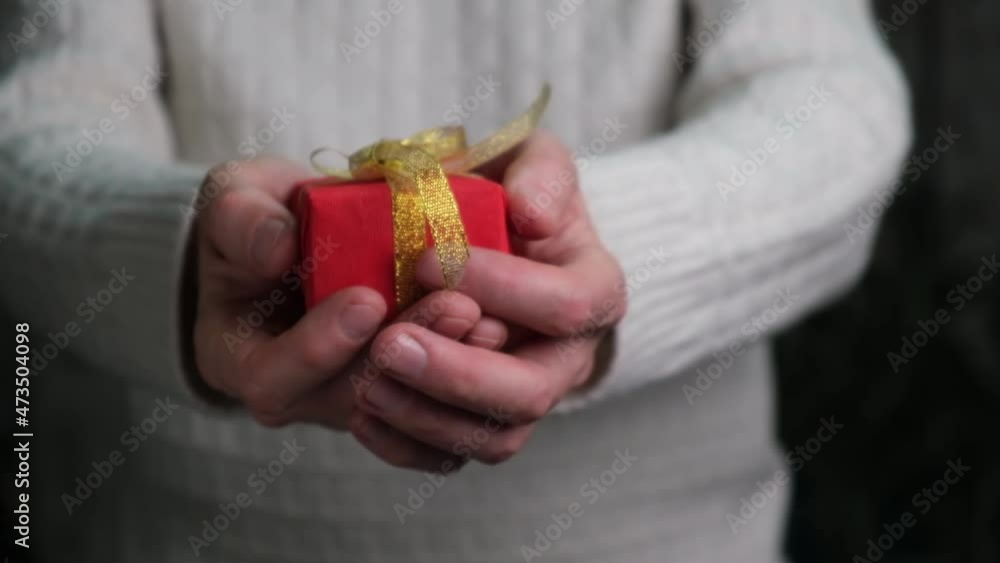 Close-up of the hands of a man in a white sweater holding out a red gift box. A man presents a gift. Elegant red gift box with a gold ribbon in men's hands. Gift giving concept. Slow motion. Close-up of the hands of a man in a white sweater holding out a red gift box. A man presents a gift. Elegant red gift box with a gold ribbon in men's hands. Gift giving concept. Slow motion.