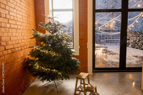 Cozy living room interior with Christmas tree in the corner. Concept of home comfort in winter. Windows overlooking a snowy garden at dusk, home with brick wall © rh2010