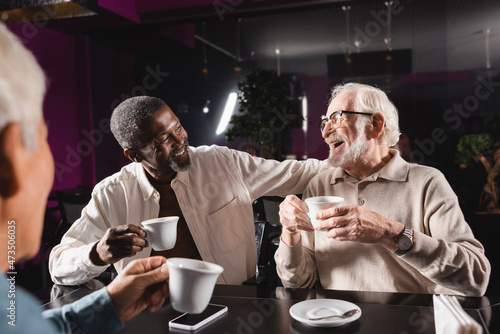 senior man in eyeglasses laughing while drinking coffee with multicultural friends in cafe