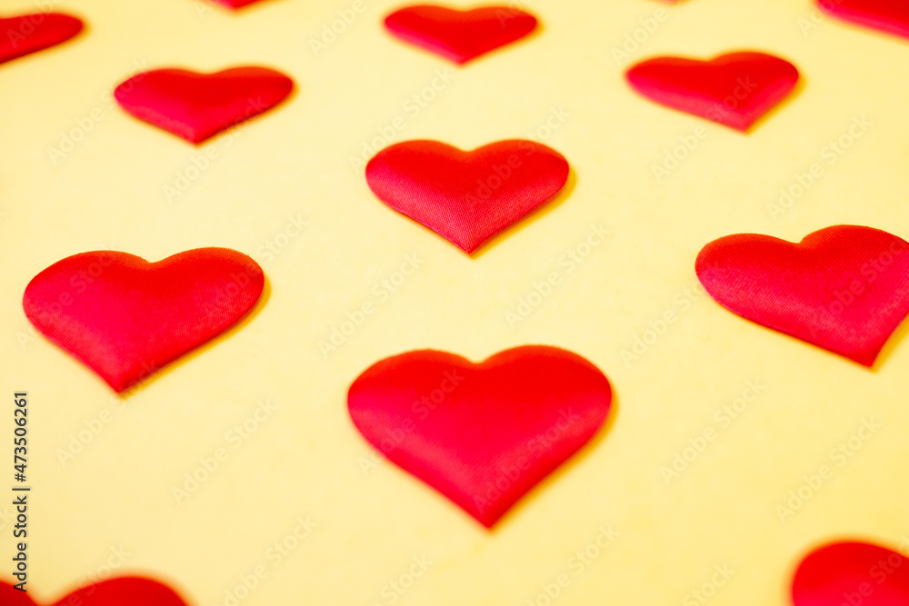 A lot of identical red silk hearts lying staggered on a yellow background. Symbol of love, tenderness and passion.