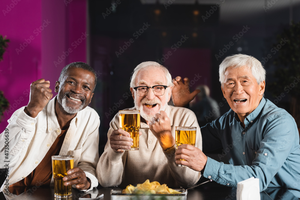 joyful senior multicultural friends with glasses of beer showing win gesture while watching football game in pub