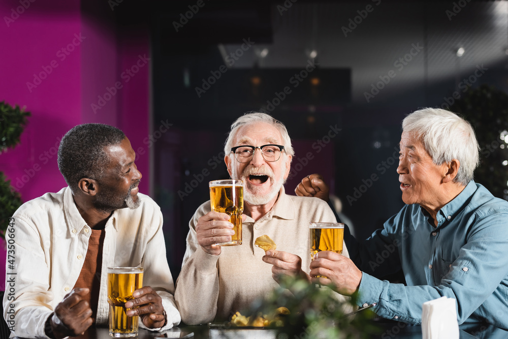cheerful man holding glass of beer near senior multiethnic friends in pub