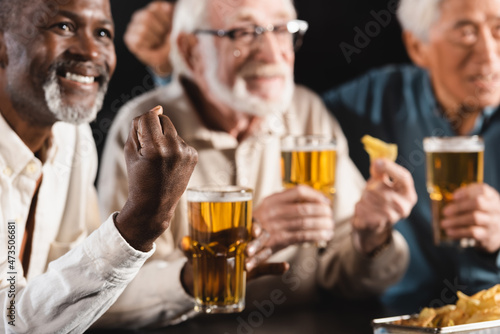 smiling african american man showing success gesture near blurred senior interracial friends watching football in pub