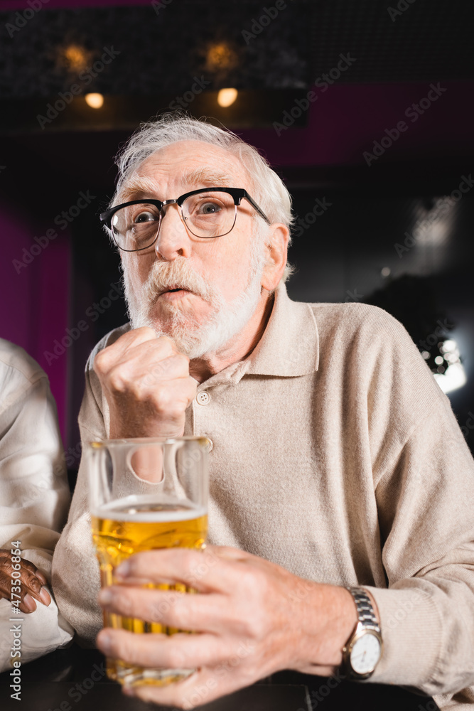 tense senior man with clenched fist and glass of beer watching football match in sports bar tense senior man with clenched fist and glass of beer watching football match in sports bar