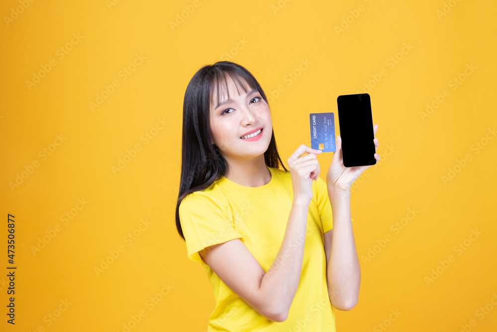 Naklejka premium Portrait Asian happy young girl smiling cheerful and showing plastic credit card while holding mobile phone isolated on yellow background