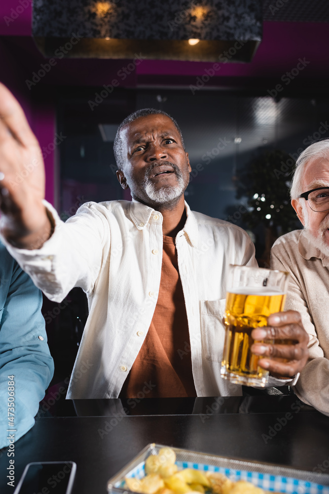 upset african american man pointing with hand while watching football match with senior friends in pub