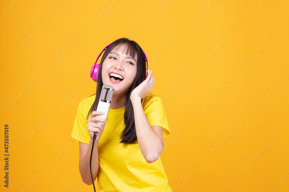 Pretty Asian woman wearing yellow shirt holding microphone singing karaoke music on yellow background. Listen music with headphones sing song in microphone. Pretty Asian woman wearing yellow shirt holding microphone singing karaoke music on yellow background. Listen music with headphones sing song in microphone.