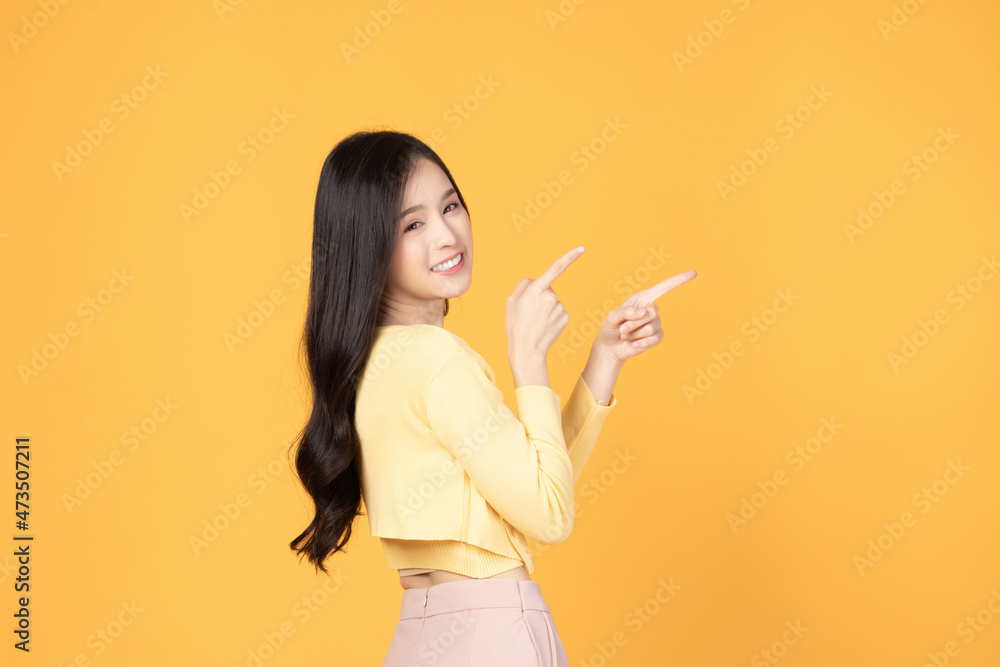 Beautiful Asian woman wearing yellow t-shirt smile and pointing finger at copy space isolated on yellow background studio portrait. Beautiful Asian woman wearing yellow t-shirt smile and pointing finger at copy space isolated on yellow background studio portrait.
