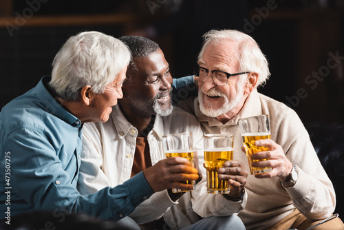 happy, elderly multiethnic friends holding glasses of beer while spending time in pub