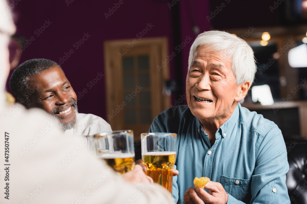 happy asian man holding beer and chips near blurred multiethnic friends in pub