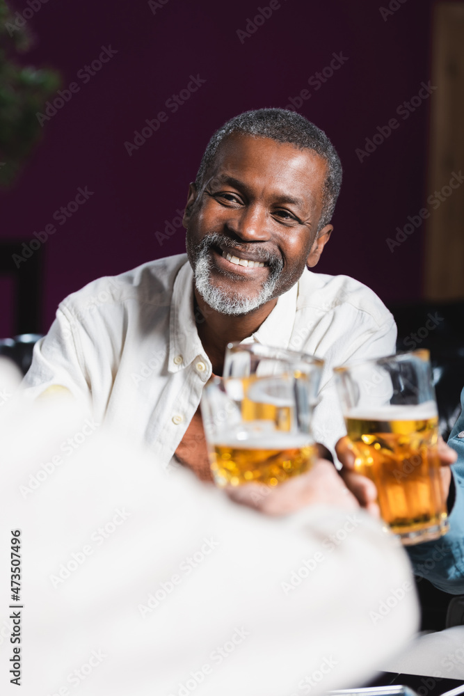 cheerful african american man clinking beer glasses with blurred senior friends in pub cheerful african american man clinking beer glasses with blurred senior friends in pub
