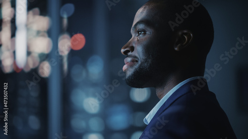 Night Office: Stylish Close-up Portrait of Powerful Black Businessman Wearing Suit Standing, Looking out of the Window on a Big City. Ambitious African CEO Thinking of e-Commerce Investment