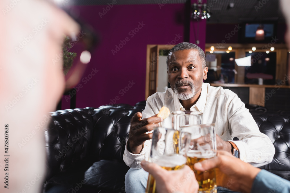surprised african american man pointing with finger while clinking beer glasses with blurred friends