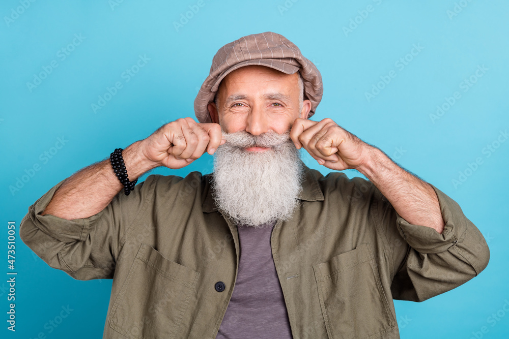 Photo of funky grey beard aged man touch mustashe wear khaki cap shirt isolated on blue color background Photo of funky grey beard aged man touch mustashe wear khaki cap shirt isolated on blue color background